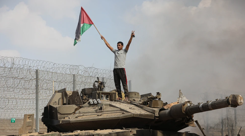 Palestinians stand next to a burning tank inside the border fence with Israel in the city of Khan Yunis in the southern Gaza Strip. Oct. 7, 2023. Photo by Yousef Mohammed/FLASH90.